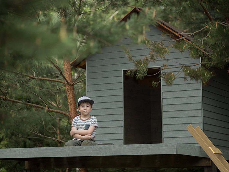 Child sitting up a tree infront of Marley Weatherboard clad tree house for JJ Roofing Supplies